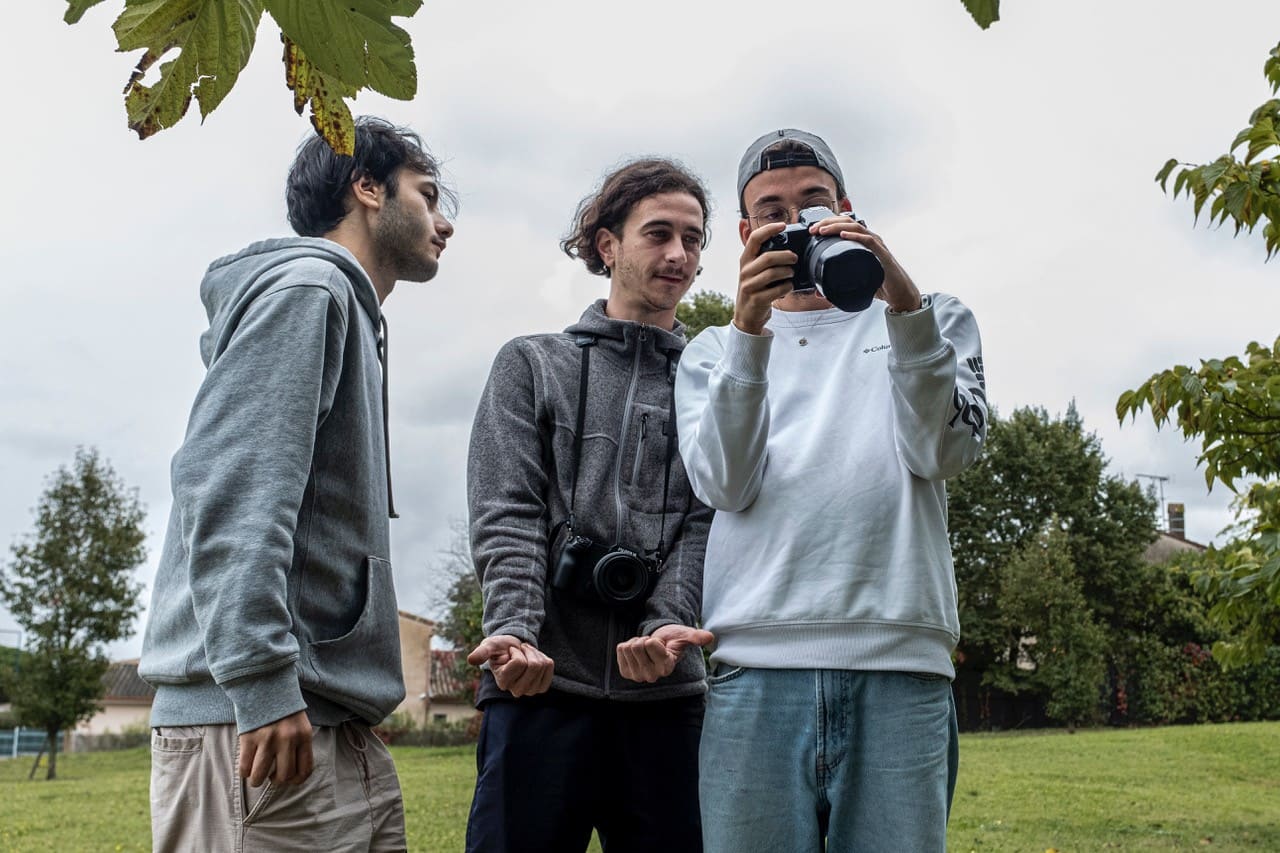 Trois étudiants en extérieur, l’un tenant un appareil photo et montrant le résultat aux deux autres, tous souriants et concentrés.