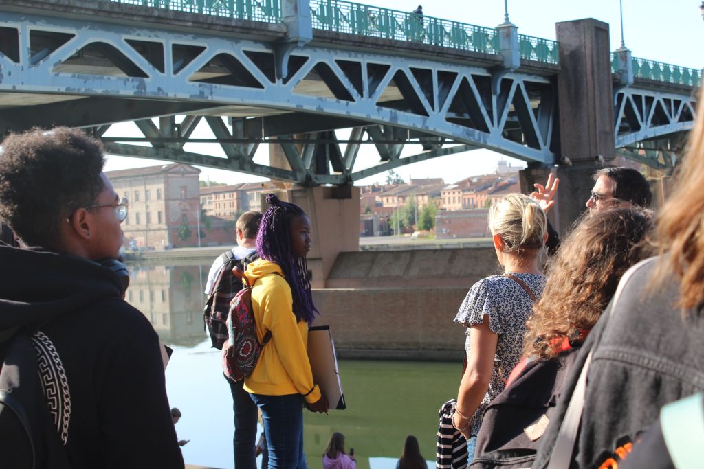 Un groupe d'étudiants participe à une sortie pédagogique près d'un pont, avec un enseignant expliquant un point précis, dans un cadre urbain ensoleillé.