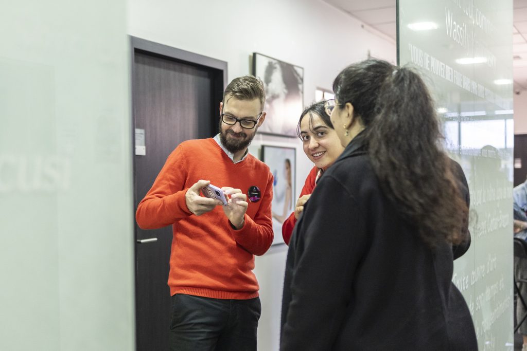 Un enseignant en pull rouge montre des images sur un appareil photo à deux étudiantes souriantes dans un couloir de l'école.