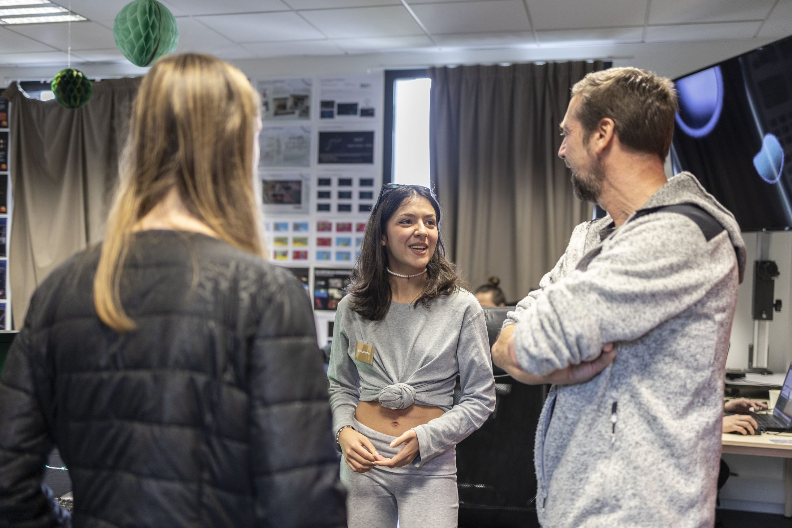 Une étudiante souriante échange avec deux membres de l’équipe pédagogique dans une salle équipée d’affichages visuels et de matériel informatique.