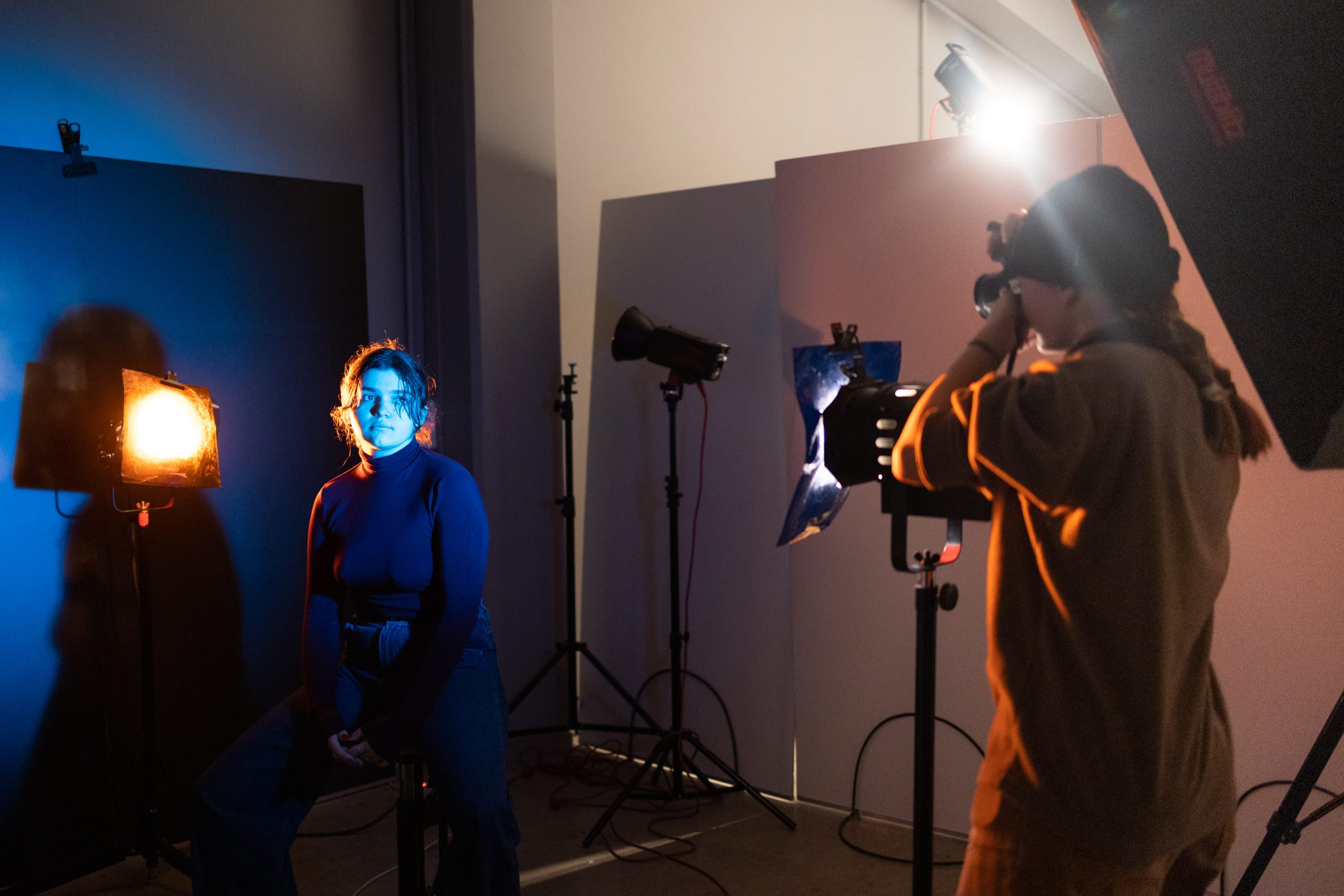 Scène de shooting en studio photo avec une lumière créative. Une étudiante prend en photo un modèle assis, éclairé par des projecteurs aux teintes orange et bleue, créant un contraste artistique.