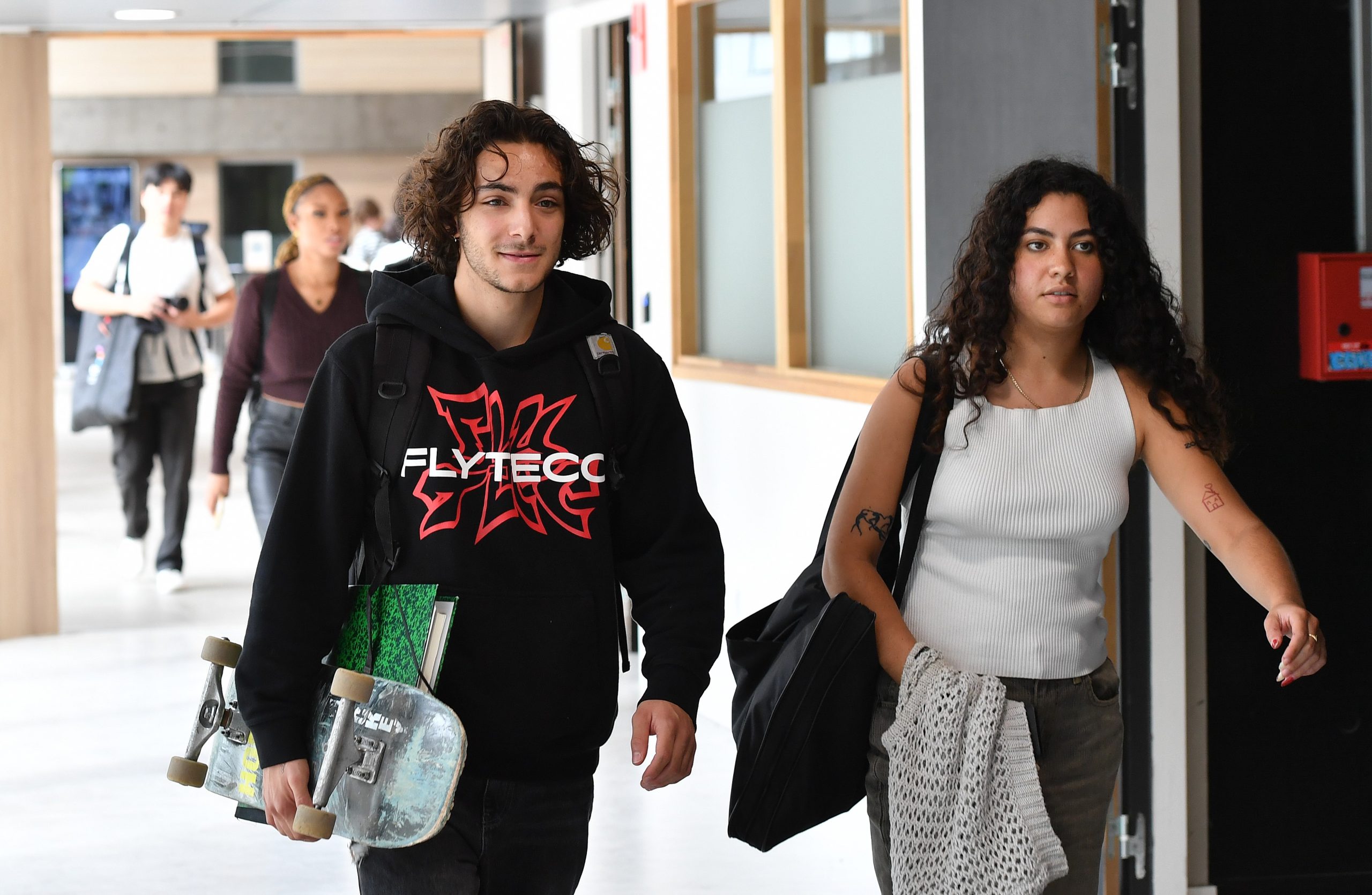 Un étudiant avec un skateboard sous le bars se dirige vers une salle de classe accompagné d'une camarade.